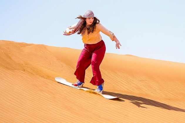 Tourist sandboarding down a tall golden dune in Dubai desert, dressed in traditional headscarf
