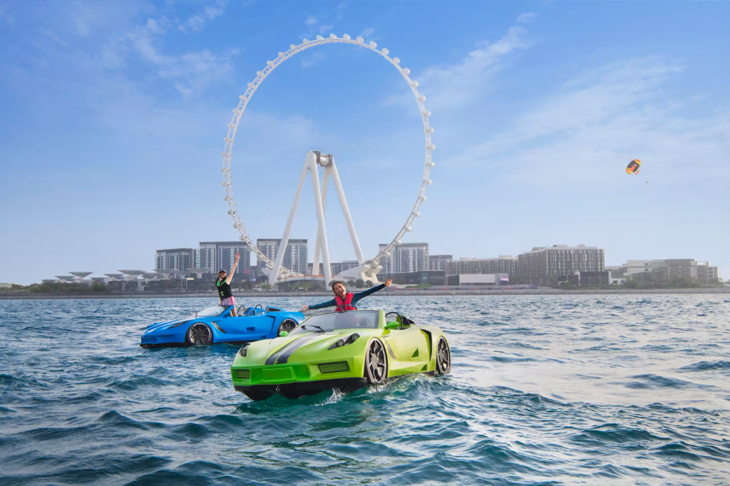 Two people riding colorful jet ski cars on the sea with Ain Dubai Ferris wheel in the background.